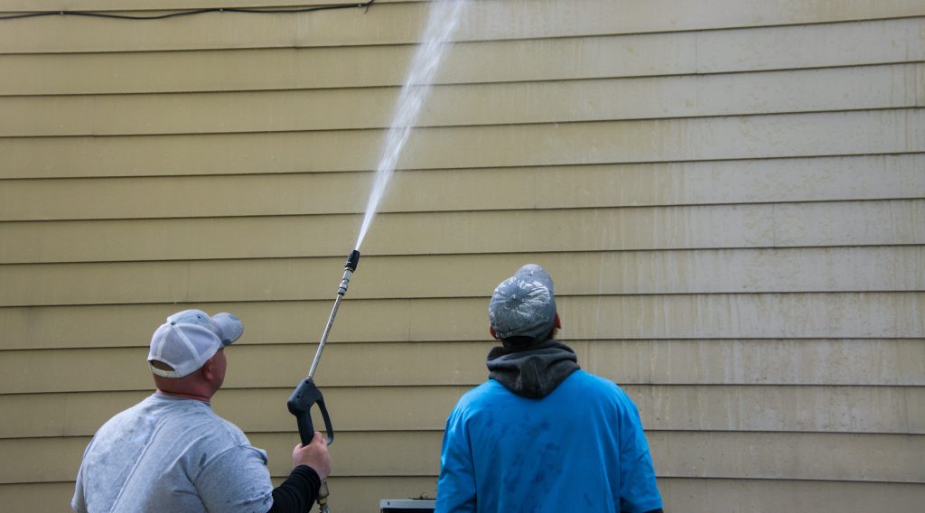 Two young men standing by the side of a yellow house. One is power washing the siding. He is holding a long nozzle and shooting the water high up on the side of the building. The other is watching; Pressure Washing