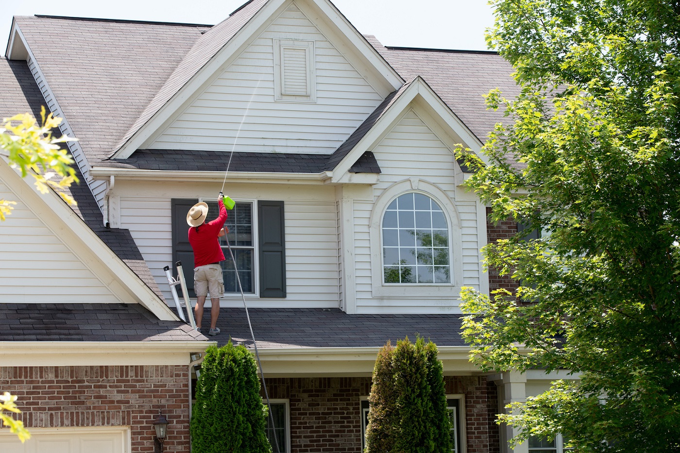 Man cleaning the upper floor exterior of his home standing on the lower roof using a pressure sprayer to wash the timber cladding and moldings on the gable