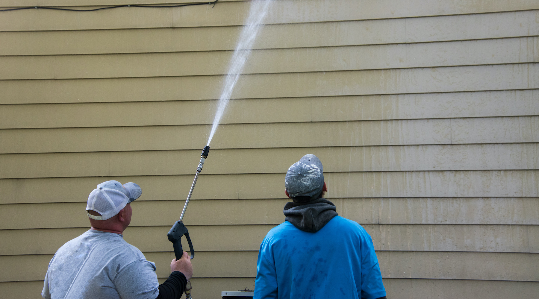 Two young men standing by the side of a yellow house. One is power washing the siding. He is holding a long nozzle and shooting the water high up on the side of the building. The other is watching